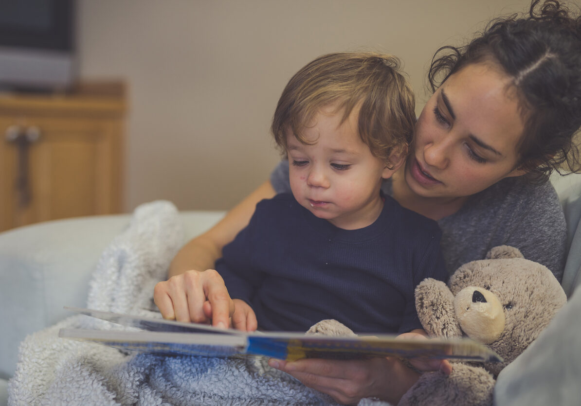 Young Hispanic mom reads a book aloud to her toddler son. They're pointing at the pictures together. A teddy bear is snuggled up with them.