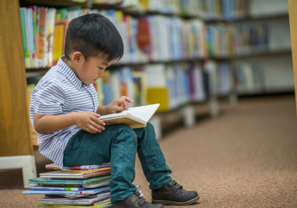 A young Asian boy is indoors in his elementary school library. He is reading a storybook while sitting on a stack of books.